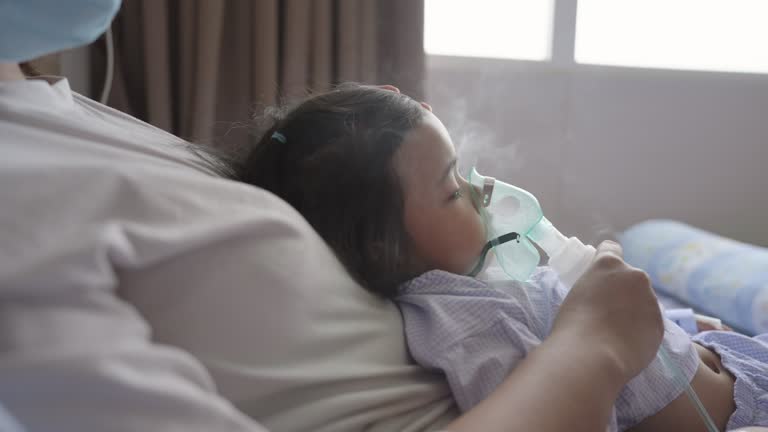 Asian mother taking care of her sick daughter sitting on a bed getting nebulizer treatment in a hospital. Asthma treatment