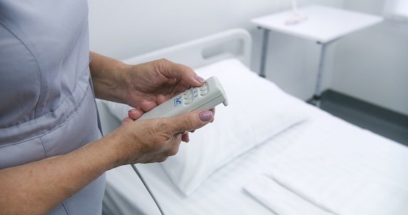 Mature nurse adjusts smart bed in hospital ward using lifting remote control. Another female medical worker prepares bedding for patient. Modern equipment in clinic or medical facility. Close up.