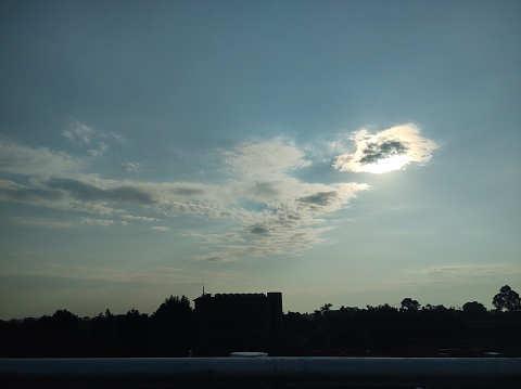 Skyscape with sun behind a cloud, buildings and trees in silhouette in the foreground