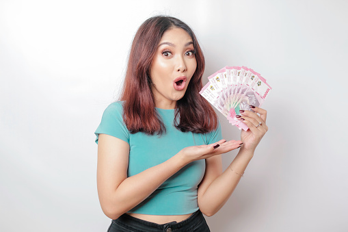 A shocked young woman is wearing blue t-shirt and holding cash money in Indonesian rupiah isolated by white background