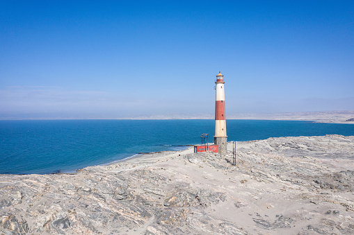 Diaz Point Lighthouse in dry desert landscape at the coast to the Atlantic Ocean under blue cloudless summer sky. Aerial Drone Point of View. The Diaz Point Lighthouse opened in 1915. It is 28 m high with a lantern and set on a one-story-high hexagonal stone base. The tower is painted in red and white bands. Located close to the City of Luderitz - Lüderitz, Karas Region, Namibia, Africa.