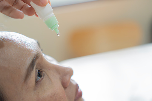 Close-up of adult Asian woman applying eye drops to treat dry eye and irritation at home. Selective focus, copy space.