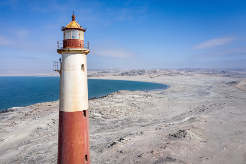 Namibia Diaz Point Lighthouse in dry desert landscape at the coast to the Atlantic Ocean under blue cloudless summer sky. Aerial Drone Point of View Lighthouse Detail View. Diaz Point Lighthouse opened in the year 1915 and is 28 m high with a lantern, set on a one-story-high hexagonal stone base. The Lighthouse tower is painted in red and white bands. Drone Architecture Detail. Located close to the City of Luderitz - Lüderitz, Karas Region, Namibia, Africa.