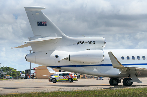 A Royal Australian Air Force Dassault Falcon 7X plane, registration A56-003, used for VIP transport of government ministers and others, is being towed to another area of Sydney Kingsford-Smith Airport. An airport security vehicle is on duty in the background.  Also in the background are Virgin Australia and Rex Airlines planes. This image was taken from Ross Smith Avenue, Mascot, behind a steel security fence, on a cloudy and sunny afternoon on 7 October 2023.