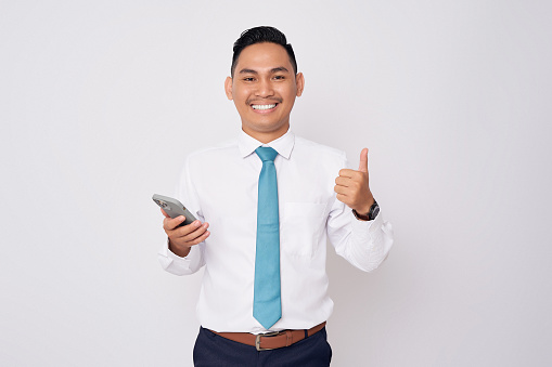 Portrait of smiling happy young Asian man in formal wear standing holding mobile phone and showing thumbs up isolated on white background