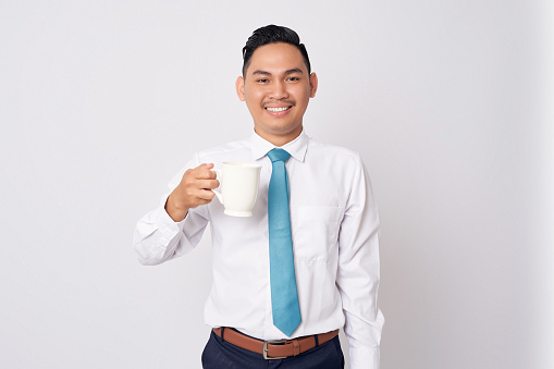 Happy smiling young Asian man in formal wear standing holding a cup of coffee, enjoying rest and looking at camera, isolated on white background
