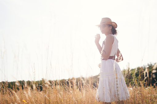 Portrait of young woman walking among high grasses in summer meadow wearing straw hat and linen dress enjoying nature. Harmony and balance.