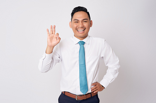 Portrait of a smiling happy young Asian man in formal wear standing confident while showing an ok sign with a finger and looking at camera with smile isolated on white background