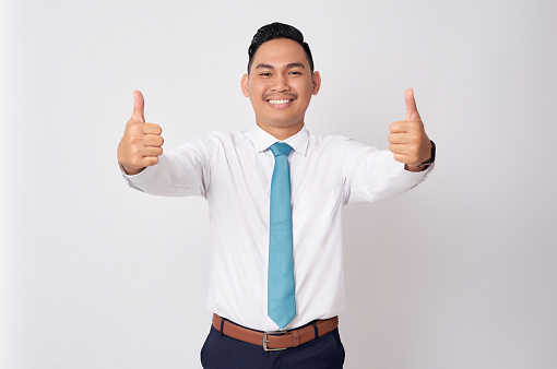 Portrait of a smiling happy successful young Asian man in formal wear standing confident while showing a thumbs up and looking at camera isolated on white background