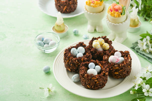 istock Easter chocolate nest cake with mini chocolate candy eggs with blossoming cherry or apple flowers on green background table. Creative recipe for Easter table with holiday decorations. Top view. 1725500591