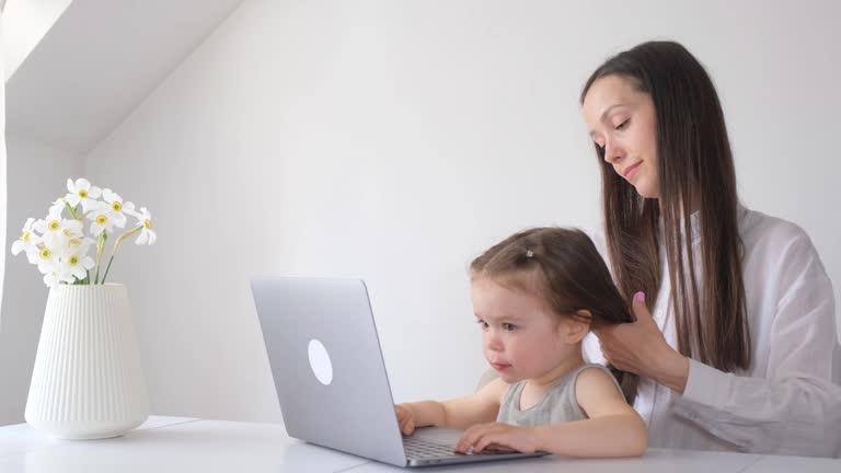 Young woman with little daughter at computer in room with modern white wall background.