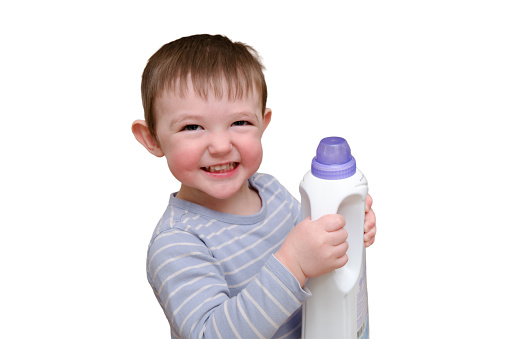 Toddler baby plays with household chemicals and detergent from the closet, isolated on white background. Child boy with detergent bottle in home living room. Kid age one year nine months