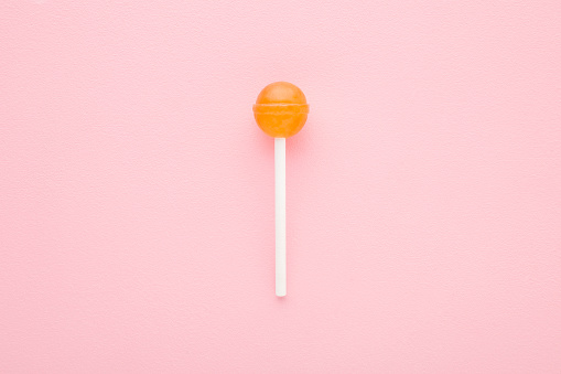 Orange lollipop on white stick on light pink table background. Pastel color. Sweet candy. Closeup. Top down view.