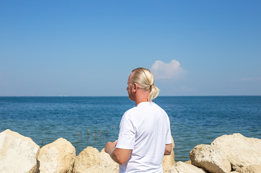 A blond man with hair in a ponytail walks along a rocky embankment, looking at the seascape. Travel and tourism.