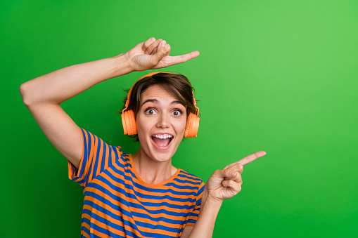 Photo portrait of lovely young lady music headphones point empty space wear trendy striped garment isolated on green color background.