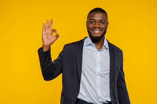 Photo of an african young happy smiling businessman dressed with elegant suit, showing ok sign with hand, isolated over yellow color background at studio.