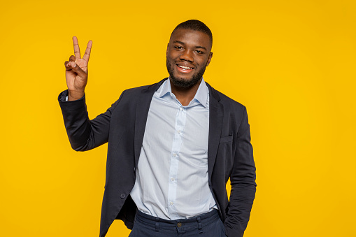 African young cheerful bearded man in business suit, does peace victory sign with hand in studio, isolated over yellow background. Businessman, business person.