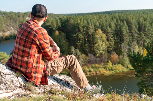 young man in cap red plaid shirt sitting in rocks and looking view of mountains covered autumn forest and river, travel and tourism landscape with copy space