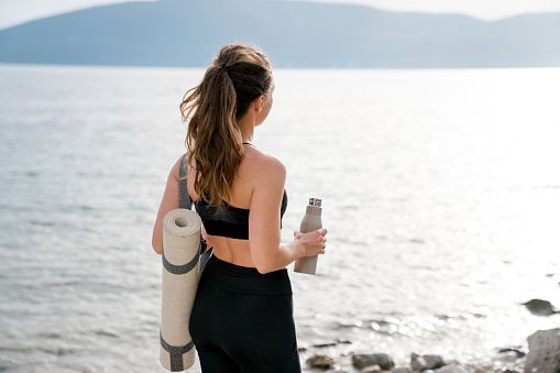 Sport, wellness on beach. Woman with yoga mat, reusable water bottle. Girl relaxing after workout outdoor, fitness in nature. Concept of healthy lifestyle, sustainable travel, ecological responsibility