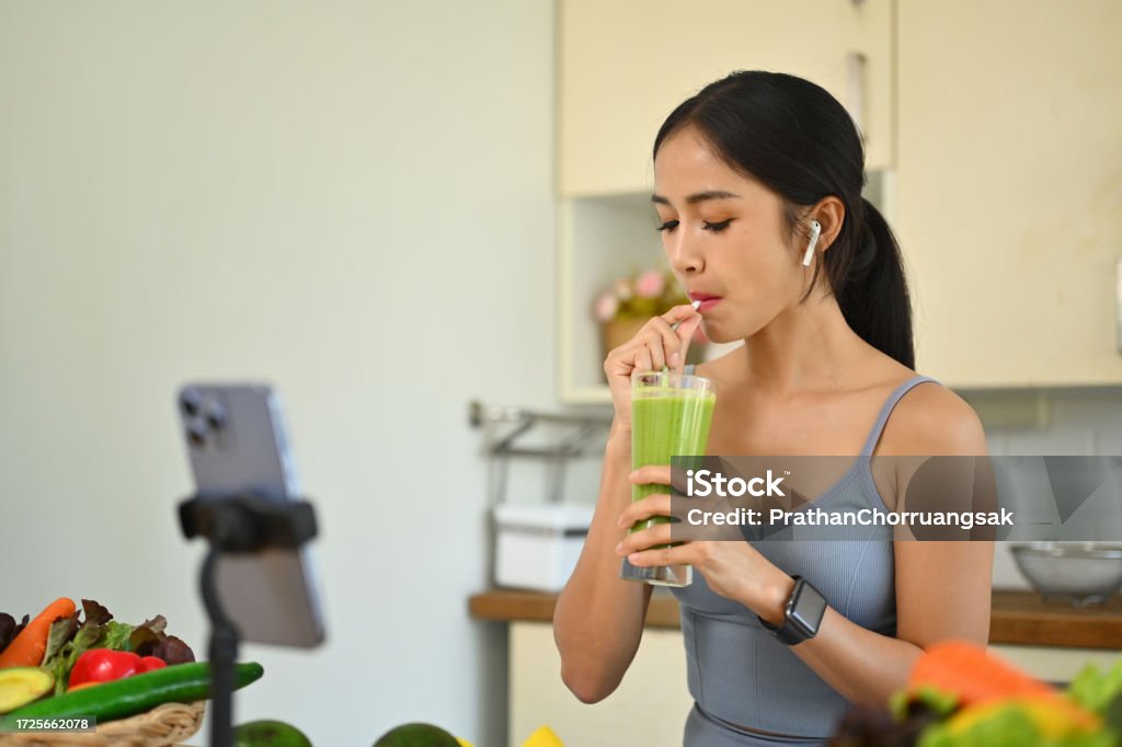 Fit young woman in sportswear drinking green vegetables smoothie while recording her blog about on smartphone Fit young woman in sportswear drinking green vegetables smoothie while recording her blog about on smartphone. 25-29 Years Stock Photo