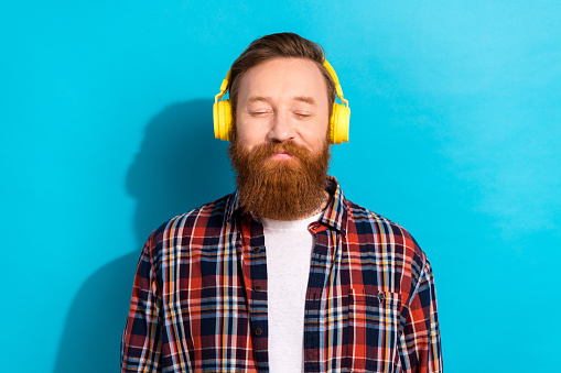 Photo of peaceful relaxed man with stylish beard dressed checkered shirt listen calm music in headphones isolated on blue color background.
