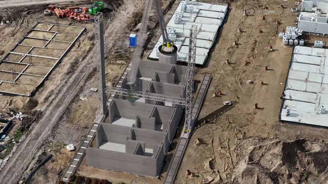 Captured in time-lapse, a large 3D printer steadily layers concrete, forming the walls of a house on a construction site.