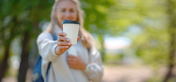 Portrait of cute woman in with cup of coffee overlooking park. Outdoor photo of relaxed female student enjoying hot drink in summer park.