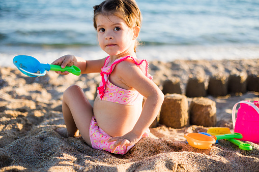 The pretty little girl is sitting on the sand and making a sandcastle. The child is using the blue toy shovel to play with the sand on the seashore.