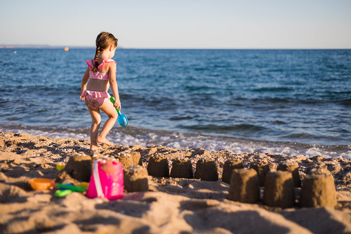 The young girl is going into the sea while holding the blue shovel in her hand. The toddler is making a sandcastle on the seashore and playing with the salt , seawater.