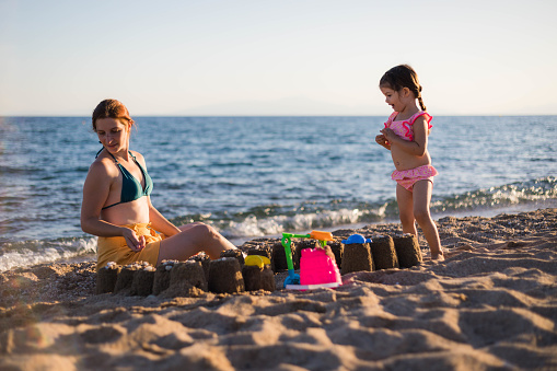 The mother is sitting on the seashore looking for stones to decorate the sandcastle they have built.