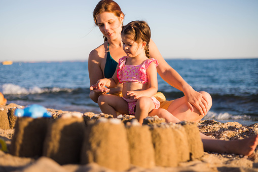 The child is sitting on her mother's lap and using some stones to put on her sandcastle. The little girl and her mother are playing on the seashore with some stones and sand.