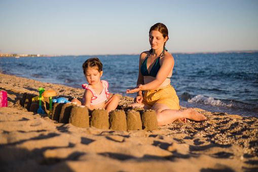 The toddler is sitting in the sandcastle she made. Her mother is giving her the colorful stones, so she can put and decorate the sandcastle.