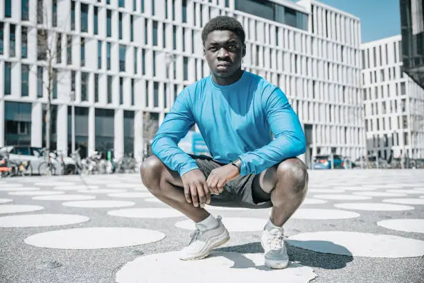portrait of young sportsman squatting on white white spotted town square  in central berlin, looking at camera