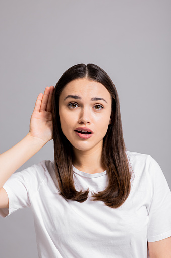 Curious surprised woman holds hand near ear listens new interesting gossip news tries to overhear stands on grey isolated background in studio wearing basic white t-shirt.