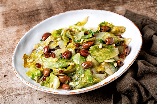 Dish with Italian salad made with sauteed broad-leaved endive or escarole with taggiasca olives, pine nuts, raisins. Vegan dish. Brown table surface.