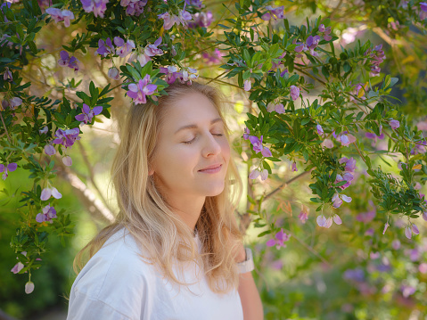 Beautiful young woman with long blonde hair and perfect skin wearing white t-shirt posing near blooming trees in garden. Nude make up. Close up portrait