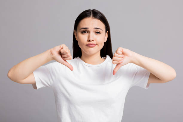 istock Tired exhausted disappointed young woman showing thumb down dislike at camera thumb down wearing casual white t-shirt on grey background in studio 1725801193