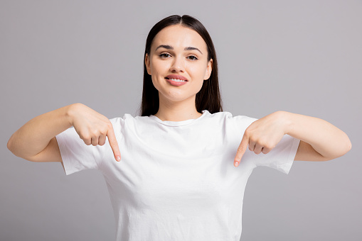 Portrait of young beautiful woman pointing fingers down smiling, showing banner, click on link below gesture, inviting people to follow, standing over white background.