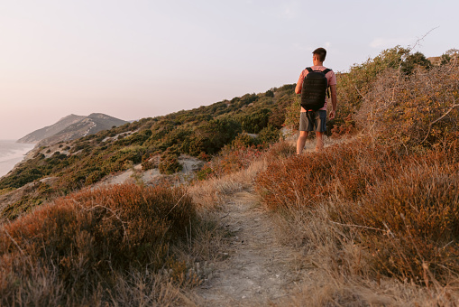 Hiker man in a T-shirt and shorts with backpack walking on the coast with warm sun light.
