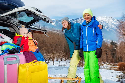 Mom stand hugging with children standing by open car trunk arrived at alpine skiing resort unloading suitcases and baggage