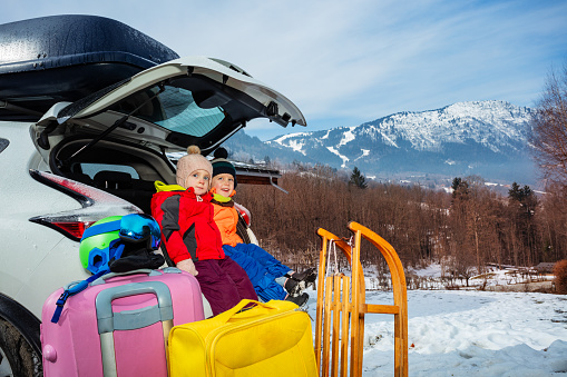 Two happy little twin kids brother and sister sit in the open car by sledge, baggage trunk ready to enjoy alpine vacation
