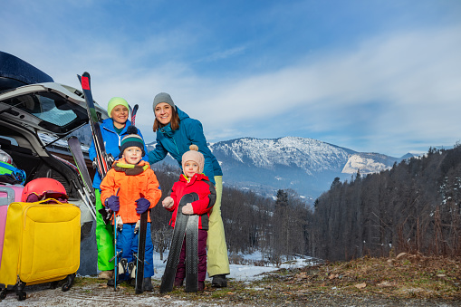 Mom stand holding ski by open car trunk arrived with 3 kids at alpine skiing resort unloading suitcases and baggage