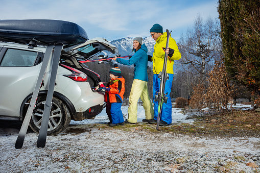 Family with mom dad and two adorable little kids standing joking unloading ski luggage by the open car trunk