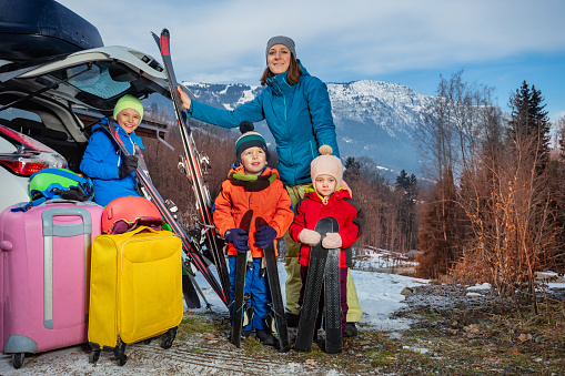 Mother stand holding ski by open car trunk arrived with three children at alpine skiing resort unloading suitcases and baggage