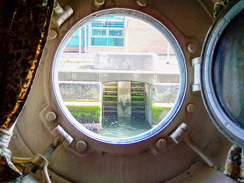 Open porthole of a very old ship looking out to a dock wall.