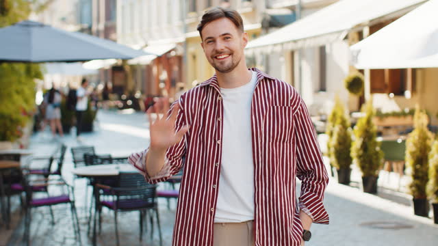 Young man smiling friendly at camera, waving hands hello, hi, greeting or goodbye in city street