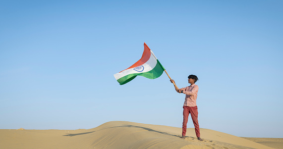 Young Indian boy is standing on the top of a sand dune and waving Indian national flag - desert village, Thar Desert, Rajasthan, India.