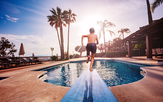 Boy run on diving board to swimming pool, sunbeds and umbrellas lit with warm sunset light going through leaves of palm tree