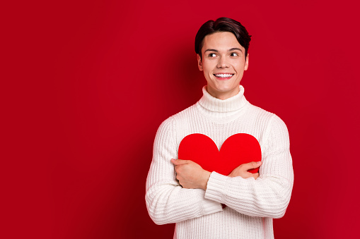 Photo portrait of dreaming boyfriend guy valentine day hold paper love heart symbol look empty space wish gift isolated on red color background.