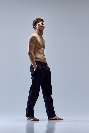 Full-length side view portrait of young man posing looking away in trousers isolated over grey studio background. Male muscular build body. Concept of natural beauty, body and skin care, masculinity.